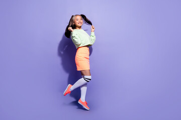 Fototapeta na wymiar Full length photo of carefree millennial miniature girl jump air touching ponytails and have some fun isolated on purple color background