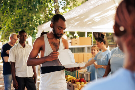Image of african american homeless guy holding his free food provided by the nonprofit group. Volunteers working to feed the needy, underprivileged and disadvantaged at an outdoor food bank.