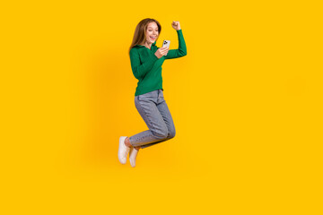 Full body length photo of girl in green pullover jumping with smartphone celebrate million followers isolated on yellow color background