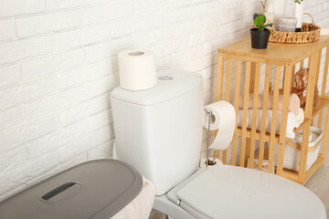 Roll of paper on toilet bowl and rack with bath supplies near light brick wall, closeup
