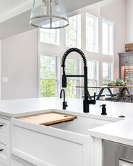 A black faucet in a white kitchen with a white farmhouse sink and chrome light hanging above the marble countertop.