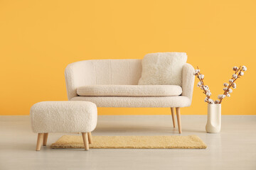 White sofa with pouf and cotton branches in vase near orange wall