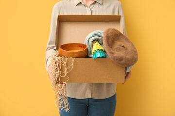 Woman holding box of unwanted stuff for yard sale on yellow background - obrazy, fototapety, plakaty