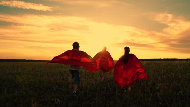 Child imagine being comic book heroes running across meadow in red capes. Children in red capes run across field pretending to be superheroes. Kids actively spend evening dressed as superheroes, dream
