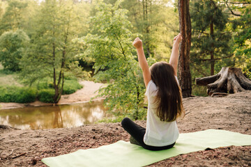 Sporty girl with long hair sitting on green mat, rising arms while enjoying nature. Child resting in front of river.