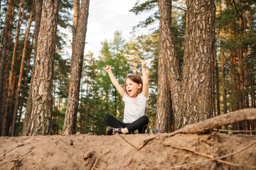 Happy female kid with open mouth sitting on ground in forest with raised arms. Sporty girl enjoying hiking in nature.