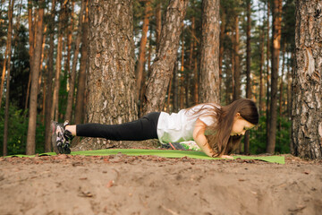 Strong little female sportsman standing on plank on green mat in nature. Focused athletic girl training outdoor.