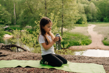 Little brunette lady calmly sit on sporty mat outdoor and hold bottle. Child look at flask resting in front of river.