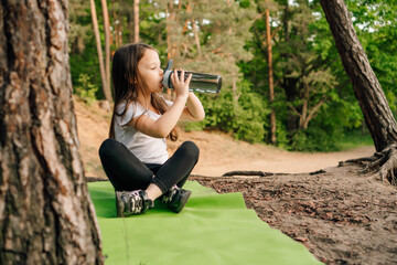 Little girl with dark long hair sitting on green rug in nature and drinking water from bottle, resting after workout.