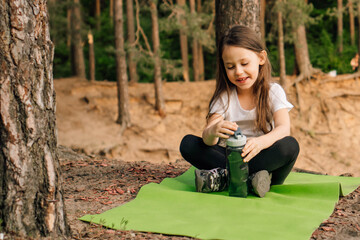 Delightful smiling female kid sitting on green sporty rug outdoor and opening bottle of water that holding in hands.