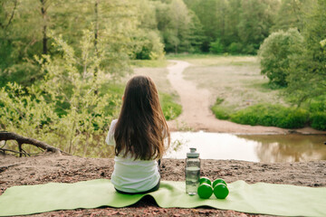 Little female sportsman sit on mat on hill in front of lake and enjoy nature. Dumbbells and bottle lying near child.