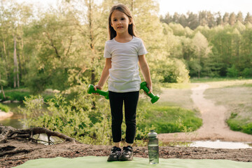 Sporty little girl holding dumbbells while standing on green mat outdoor. Junior athlete enjoy training on warm day.