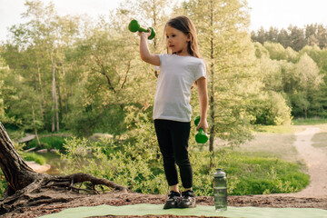 Dynamic little sportsman working out with dumbbells in hands on sporty mat outdoor. Girl lifting weights in nature.