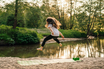 Active little athlete with long hair jumping high on sporty rug on sandy beach near lake. Girl training in nature.