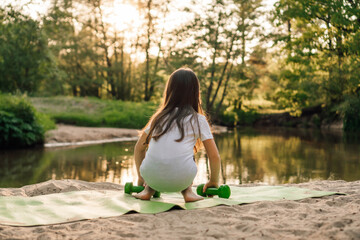 Little girl with small dumbbells in hands squatting down on green mat while workout on sandy beach in front of river.