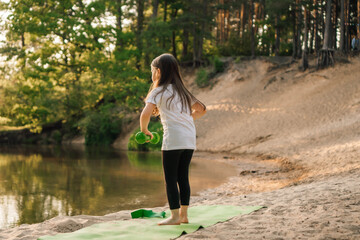 Junior female athlete bent down to put small green dumbbells on sporty mat. Little girl training on beach near river.