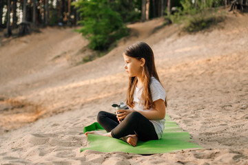 Little lady sit on green sporty mat on sand and hold bottle of water. Girl look at flask, dumbbells lying near kid.