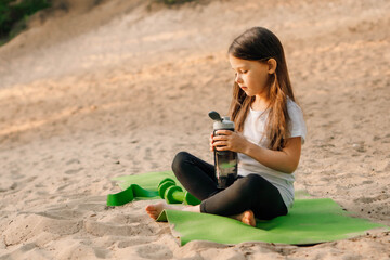 Little lady sit on green sporty mat on sand and hold bottle of water. Girl look at flask, dumbbells lying near kid.