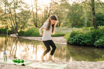 Delightful little female athlete happily jumping on green mat on sandy beach near river. Girl doing sports outdoors.