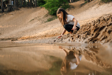 Curious girl with brown hair squatting down on sand near river in forest and going to take grit....