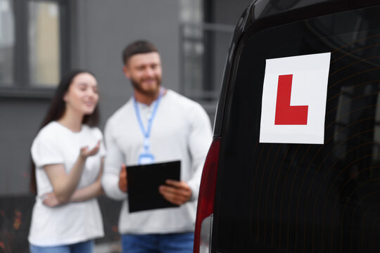 Learner driver and instructor with clipboard near car outdoors, selective focus on L-plate. Driving school