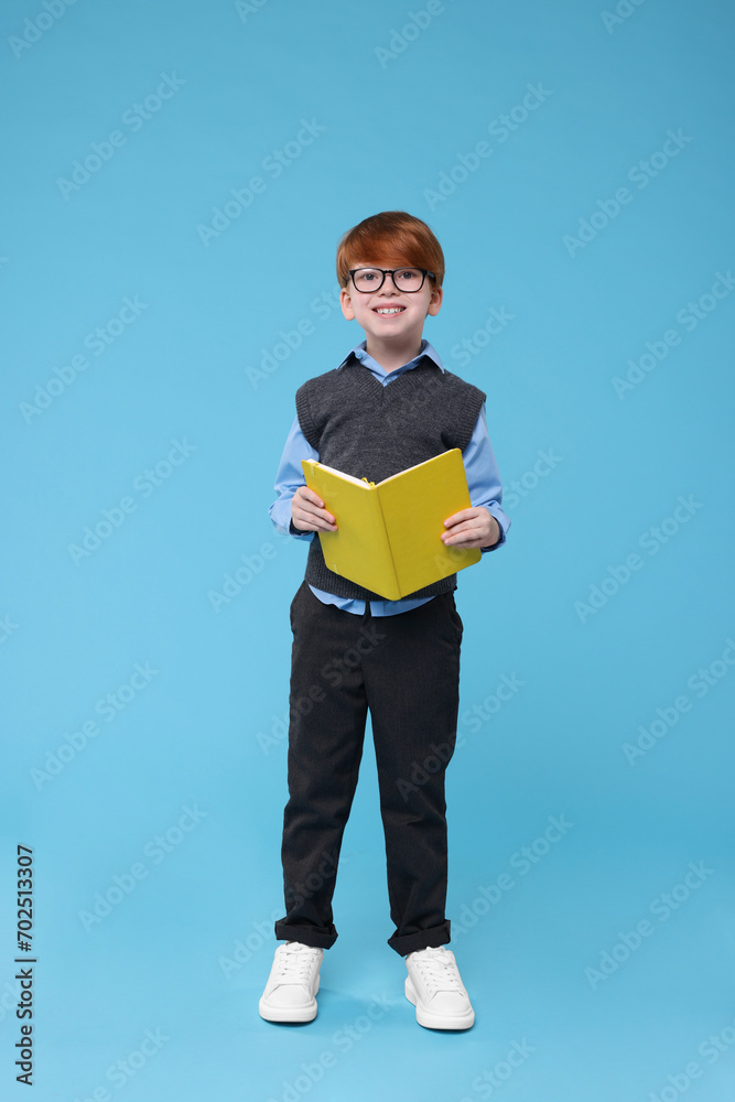 Wall mural Smiling schoolboy in glasses with book on light blue background