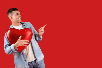 Handsome young man with heart-shaped air balloon pointing at something on red background....
