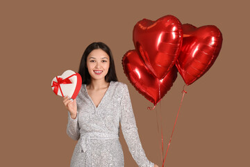Young Asian woman with heart-shaped air balloons and gift for Valentine's day on brown background