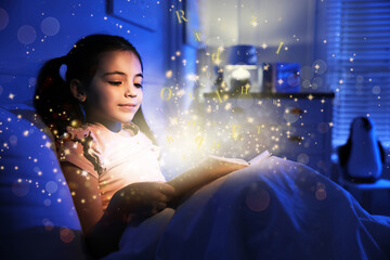 Girl reading shiny magic book with letters flying over it in dark bedroom