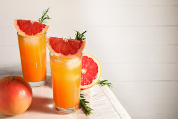 Tasty grapefruit drink with ice in glasses, rosemary and fresh fruits on light wooden table. Space for text