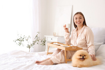 Young woman with cute Pomeranian dog having breakfast in bedroom