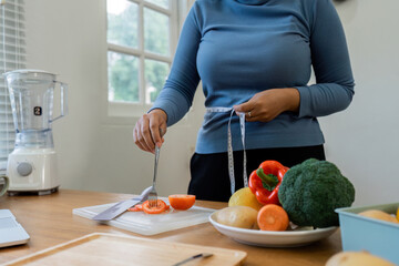 Overweight woman measuring her hip with tape measure while eating vegetable - obrazy, fototapety, plakaty