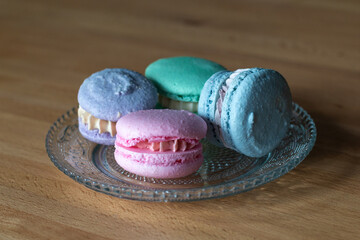 Four French macarons; pink, green, blue, and purple. The assorted elegant sandwich-style sweet dessert is stacked on a blue plate and wooden table. The traditional French pastry has a hard shell.