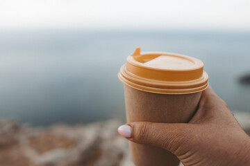 Hand holding Yellow cup with lid, coffee against a backdrop of a blue sky and sea. Illustrating cup and beverage