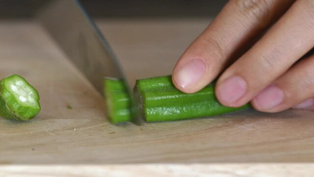 Hand holding knife and cutting okra on wooden board