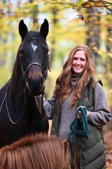 Woman with red hair and a black horse stands in the autumn forest and laughs at the camera,...
