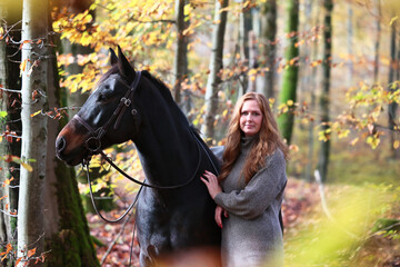 Woman with red hair in a knitted dress stands with her horse in an autumn forest, horse looks...