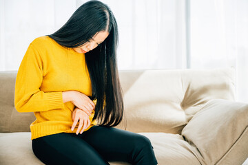 Highlighting stomachache, Asian woman on home couch endures abdominal pain. Depicting discomfort...
