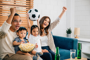 Delighted parents and children enjoy a football match at home, celebrating a goal with popcorn and...