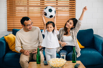 A jubilant family, with popcorn and a ball, raises their arms, cheering while watching a football...
