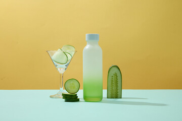 A blank plastic bottle, cocktail glass, and fresh cucumber slices adorn a pastel background-perfect mockups for imaginative advertising. Front view.