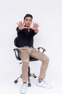 A worried an annoyed young man sitting on a chair saying no, gesturing to stay away with both hands. Not wanting to take responsibility. Isolated on a white background.