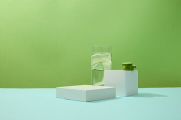 A glass of detox water with thin slices of cucumber and two white platforms displayed on a pastel...