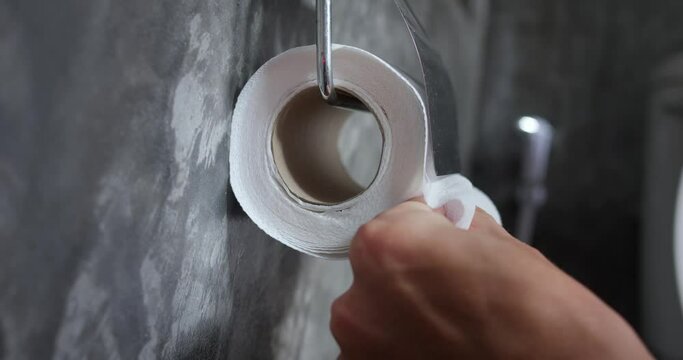 Hand tears off toilet paper in holder. Sensible use of toilet paper, reasonable consumption, tearing off sufficient amount. Grey wall on which holder located, bathroom, toilet room. WC, male man hand