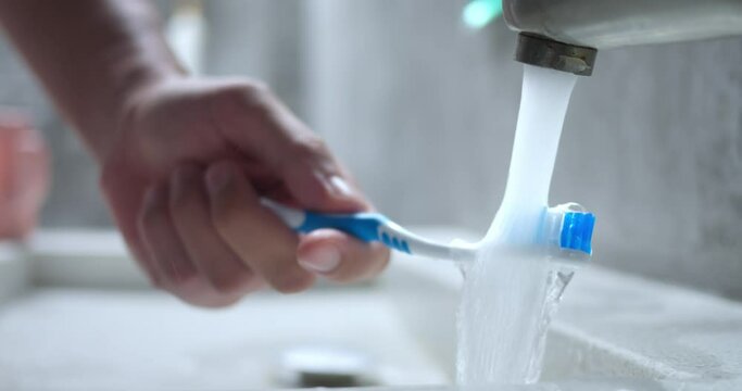 Hand rinsing toothbrush under water. Close up, water flowing in sink from tap, blue brush rinsed under running water to soften bristles and further brush teeth. Man male hand holding toothbrush.