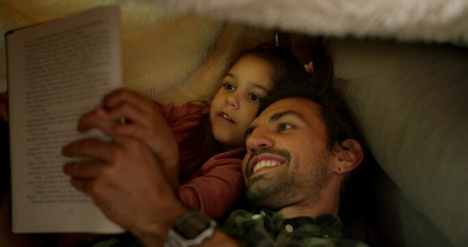 A little brunette girl in a pink dress lies in bed with her father who tells her stories, they read a book together and look at pictures with a flashlight on near a gray sofa at home