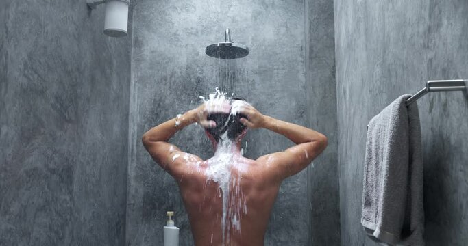 Man washing in shower back view. Man soaping foaming hair, washing hair standing under shower in grey minimalist small shower room. Taking shower, maintaining hygiene cleanliness of body. Slow motion