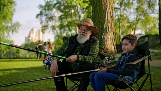 Grandfather and kid boy spending weekend together. Grandpa talking with grandson in summer day at the river. Family fishing together. Concept of grandparent speaking to grandchild holding fish rod