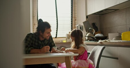 Shooting from behind the wall, a happy man in a green checkered shirt is sitting at the kitchen table with his little daughter, a brunette girl in a pink dress, who is doing a manicure for her father