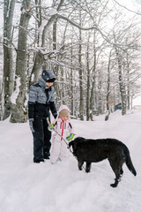 Little girl hands a stick to a black dog while standing with her mother in a snowy forest